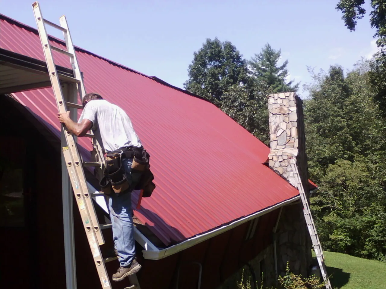 Roofer on a ladder beside a red metal roof and stone chimney — Soffit & Fascia Repair by Roofing Brothers in Lanesborough
